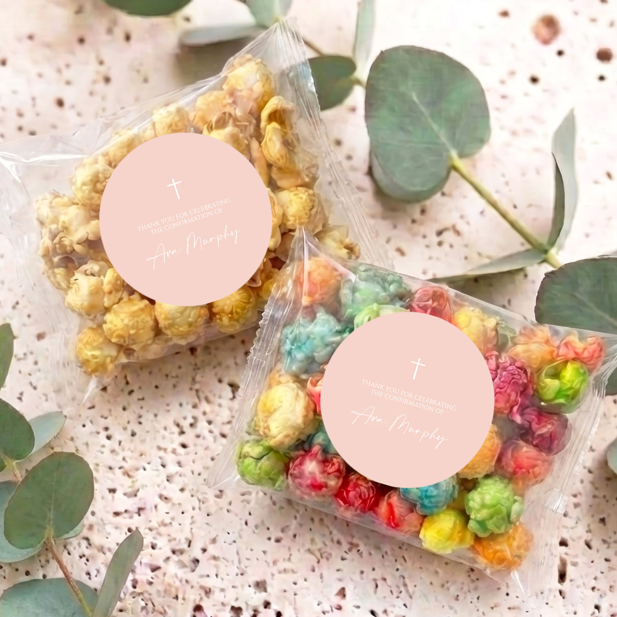 Two clear popcorn treat bags with pastel popcorn and peach round labels featuring a cross