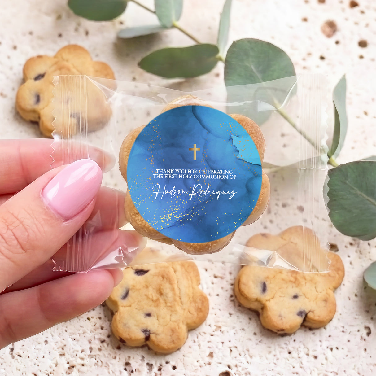 Hand holds bite-size chocolate chip cookie with blue First Communion gold cross label