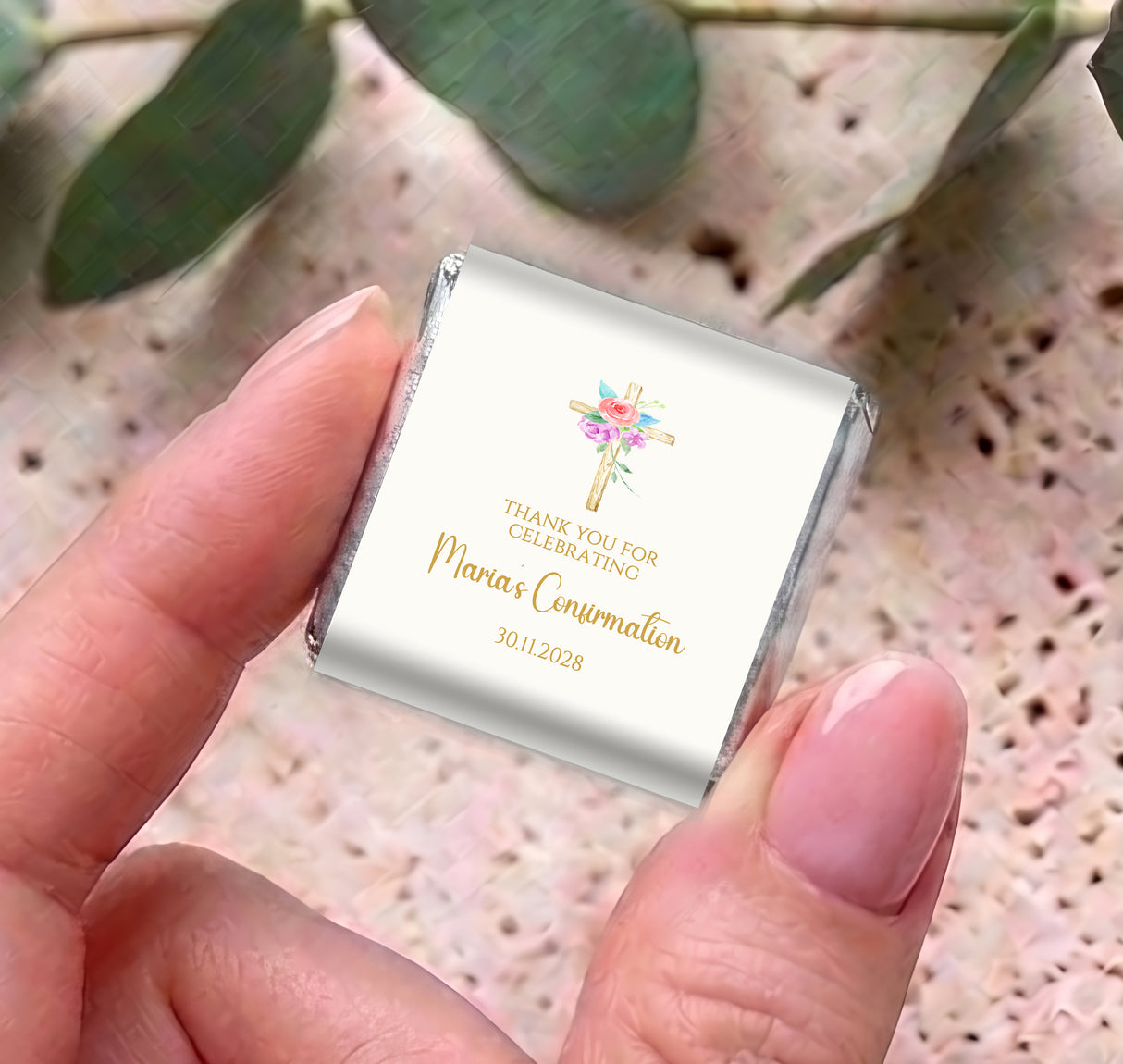 Personalized mini chocolate favor held in hand, floral cross and gold text on silver wrap