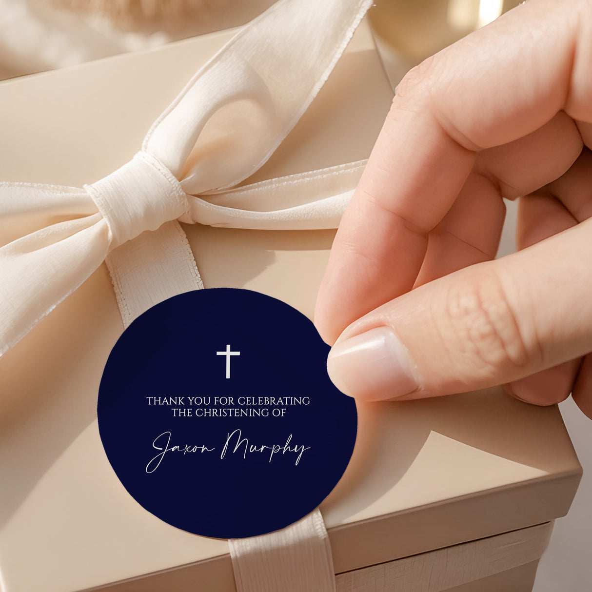 Round navy christening label with white cross, text, and name on cream ribbon