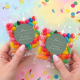 Two clear jelly bean favor bags with colorful beans and sage green 'Love' round labels