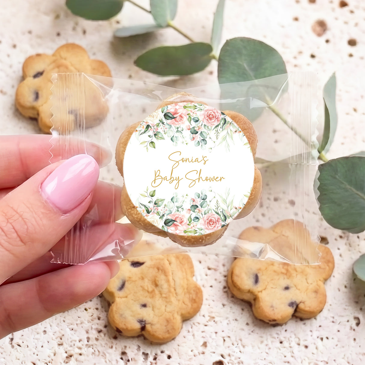 Hand holding a cookie with floral "Sonia's Baby Shower" round topper and clear bag