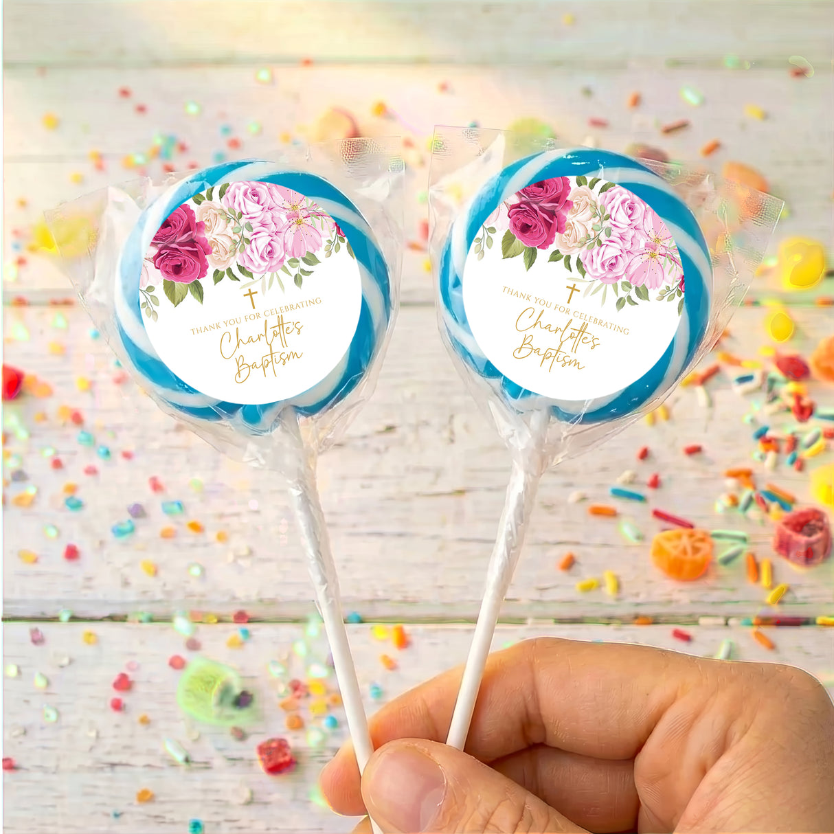 Two Christening lollipops in clear wrappers, held by a hand; blue rose floral design