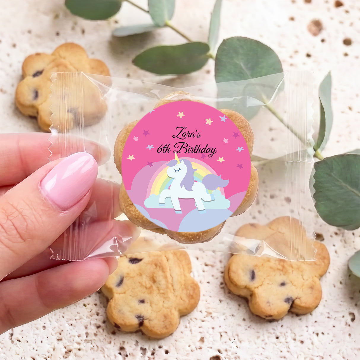 Hand holding choc chip cookie with pink unicorn birthday sticker and clear wrapper