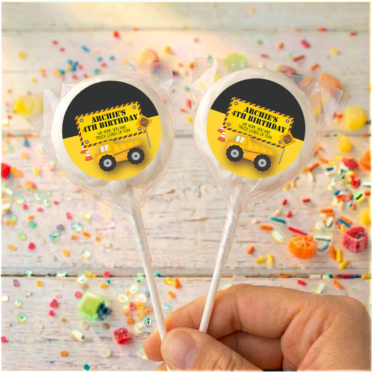 Personalised birthday lollipops with yellow truck labels on clear wrappers, held in hand