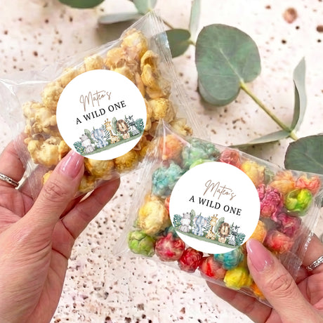 Hand holding two personalized popcorn bags, circular 'A WILD ONE' labels, mixed-color popcorn