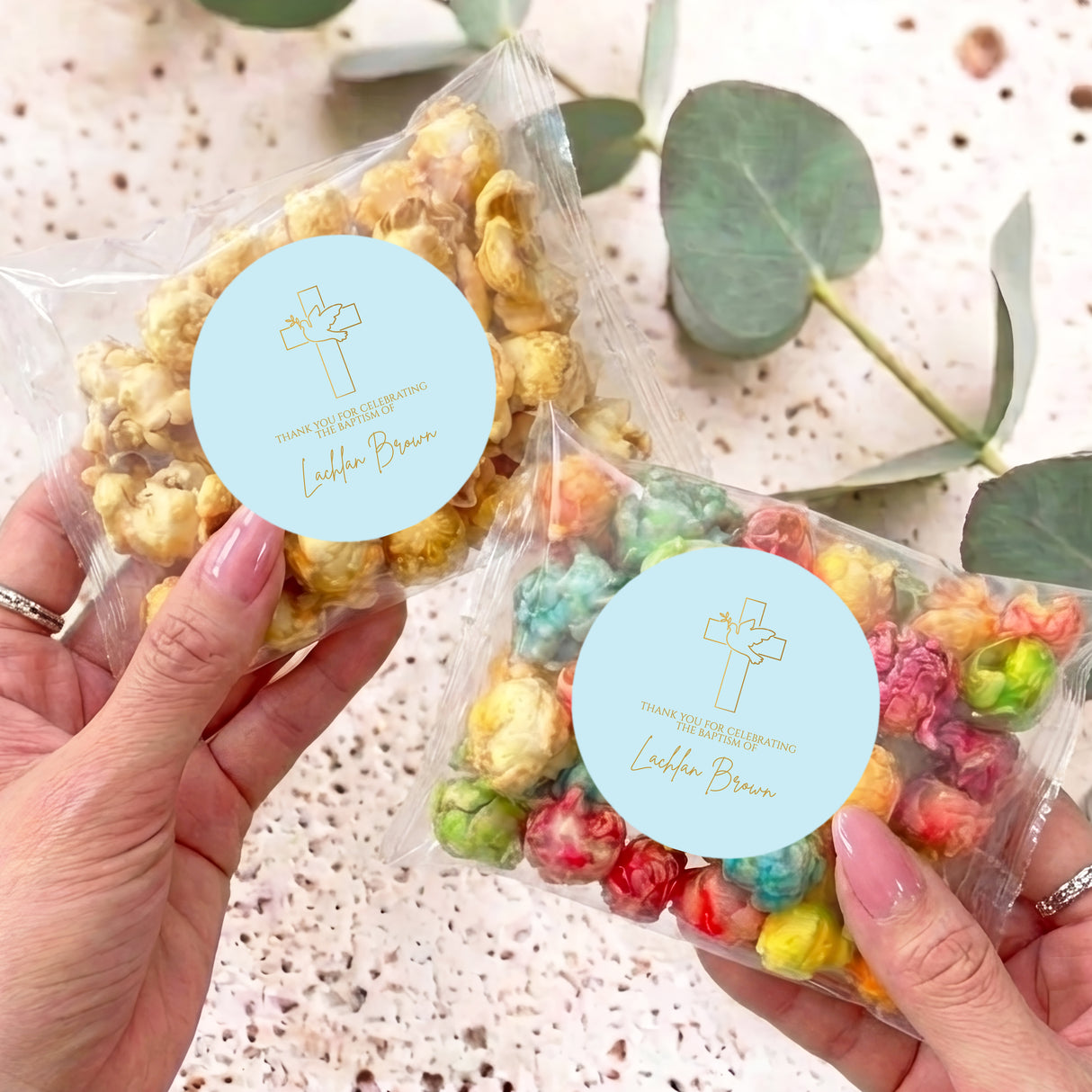 Personalised christening popcorn bag with cross label, held in hands, clear wrap.