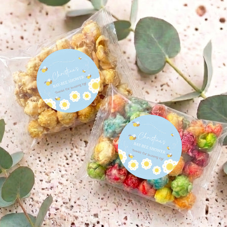Two personalised blue bee baby shower popcorn bags with clear wrap and pastel popcorn