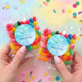 Two personalised jelly bean bags with blue labels, gold cross, held in hands, filled with candy