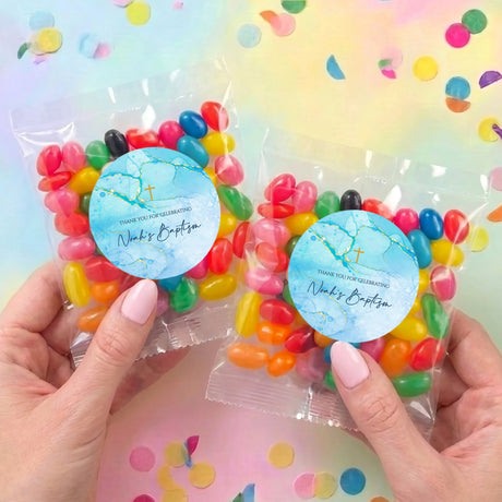 Two personalised jelly bean bags with blue labels, gold cross, held in hands, filled with candy