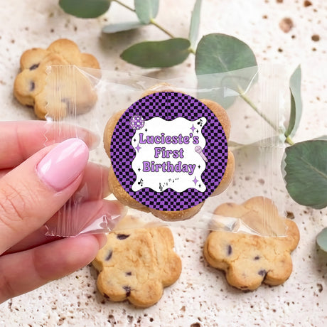 Hand holding a cookie bag with purple “First Birthday” round sticker; choc chip cookies