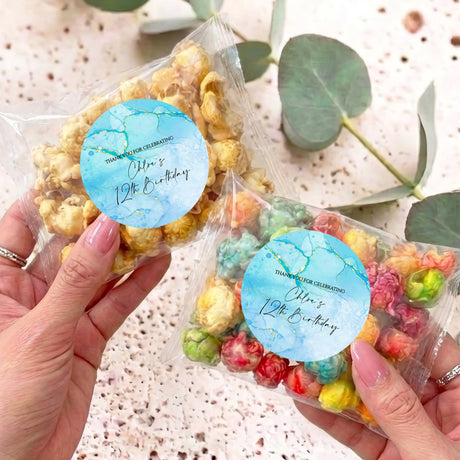 Personalised popcorn bag with blue round label and clear wrap, held in hands.
