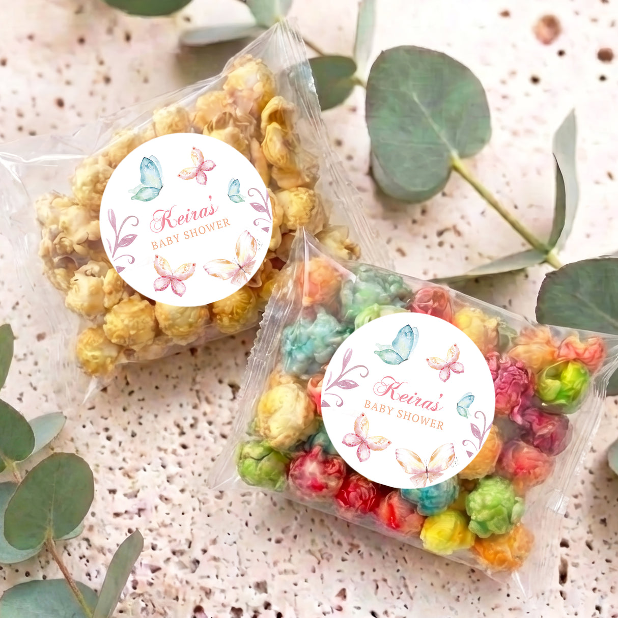 Two personalised baby shower popcorn bags with butterfly round labels, pastel popcorn pieces