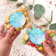 Personalised popcorn bag with clear wrapper and blue round label, held in hand.