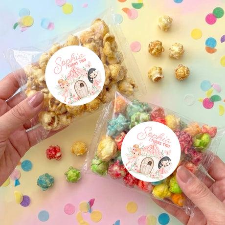 Two clear popcorn bags held in hands, each with round personalized label and mixed popcorn.