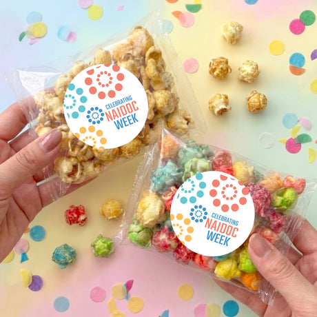 Popcorn bags in hands: two clear bags with pastel confetti and Celebrating NAIDOC Week stickers.