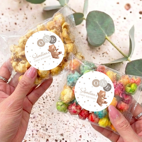 Personalised popcorn bags held in hands, clear wrap, rainbow popcorn, round 'ONE' bear labels
