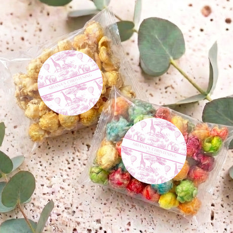 Pink safari-themed popcorn bags with round white personalised labels, lying on speckled surface