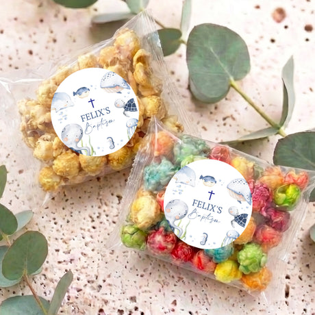 Two personalised popcorn bags with round baptism labels and sea-animal designs in clear wraps