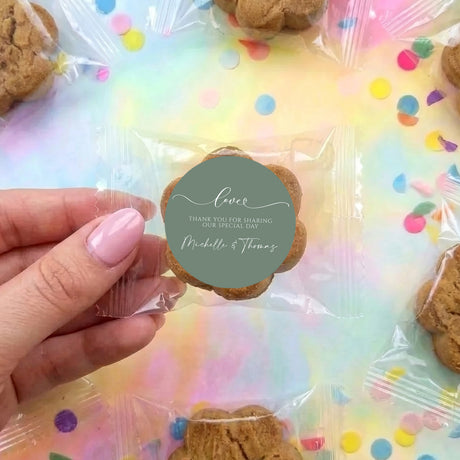 Handheld chocolate chip cookie with sage green round wedding label on clear bag