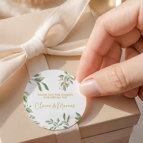 Round wedding favor label with eucalyptus leaves and gold text, held by fingers