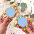 Clear popcorn bag with beige kernels and blue round personalized label, held by hand