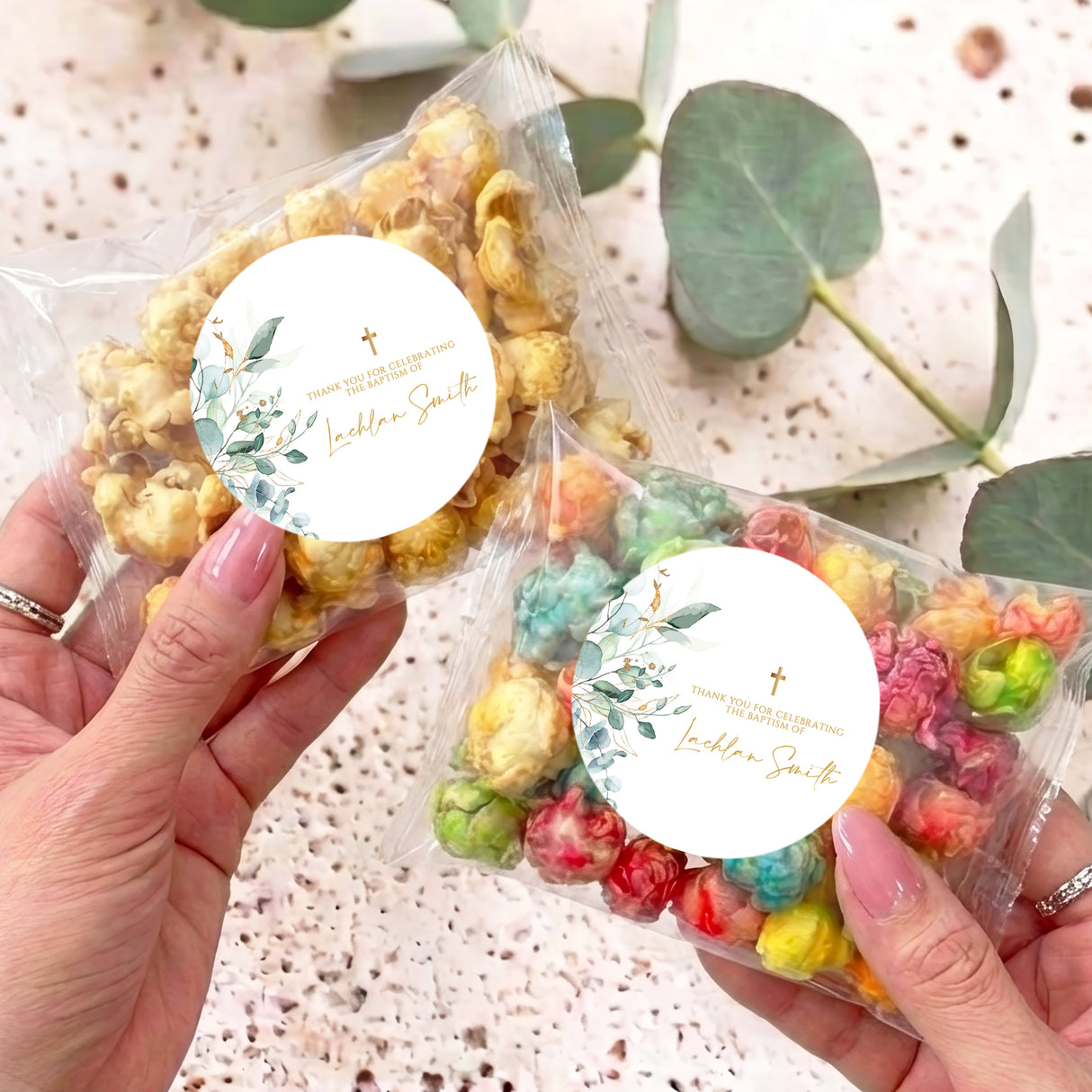 Personalised popcorn bags with round leaf-and-gold cross labels, held in hands