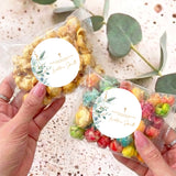 Personalised popcorn bags with round leaf-and-gold cross labels, held in hands