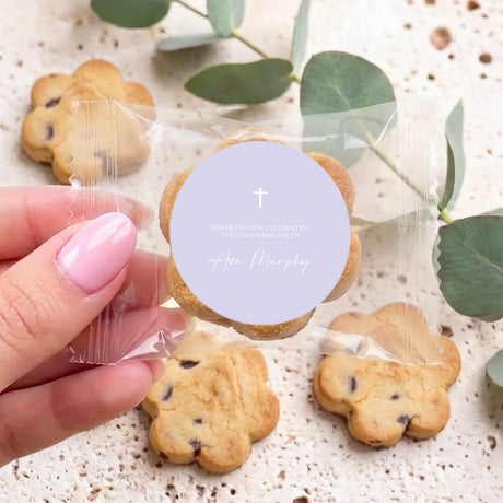 Bite-size choc chip cookie with lavender round label, cross and name text; held by hand.