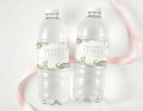 Personalised dinosaur water bottle labels on two clear plastic bottles, text 'SEBASTION IS THREE'