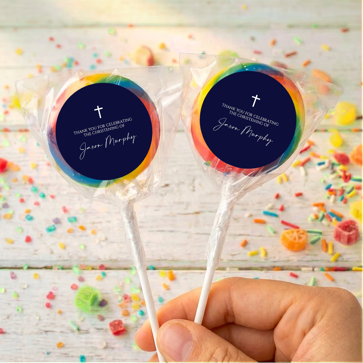 Personalized lollipops with navy round labels, white cross, rainbow halo on wrappers.