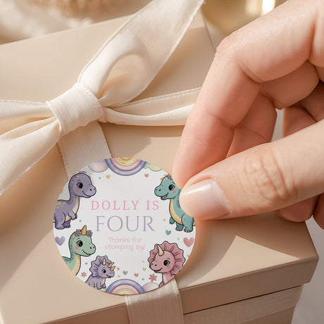 Round dinosaur birthday label with 'DOLLY IS FOUR' text and pastel dinosaurs