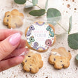Hand holding a dinosaur-themed personalized cookie with round topper reading “Dolly is four”.