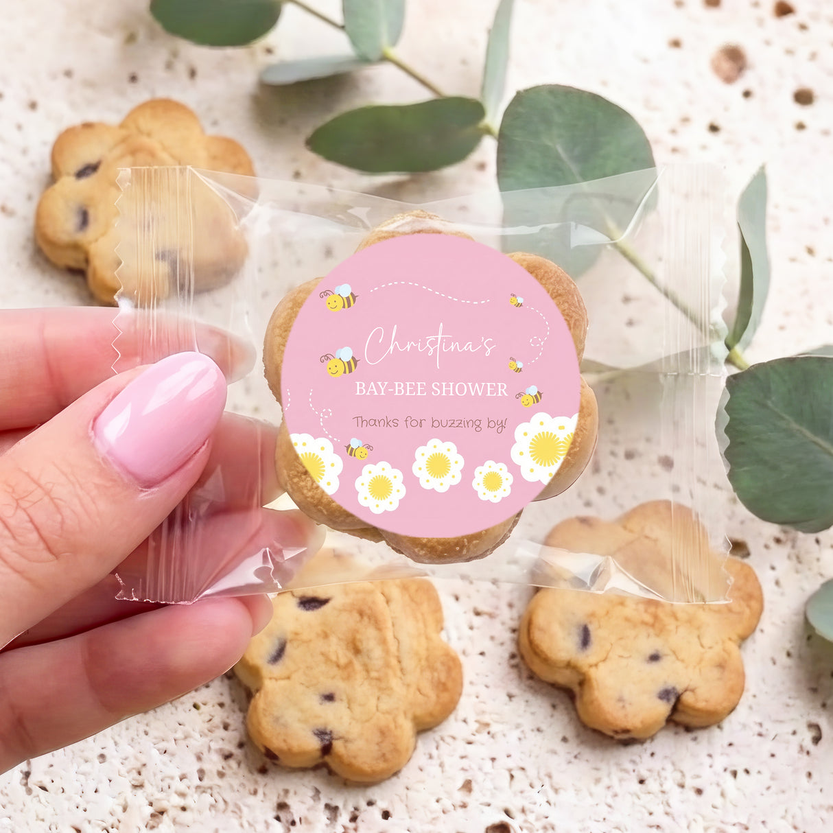 Hand holding bite-size choc chip cookie with pink 'Baby-Bee Shower' round label