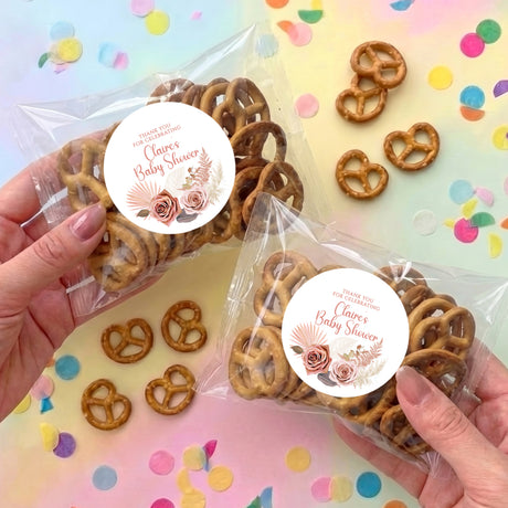 Two clear pretzel bags with brown pretzels and pink floral baby shower round labels