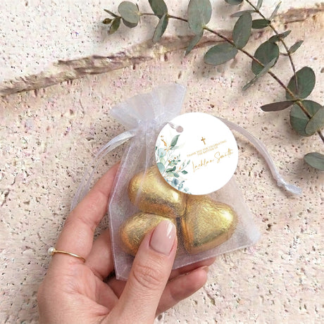 Gold organza baptism gift bag with metallic gold heart-shaped chocolates held by hand and round tag