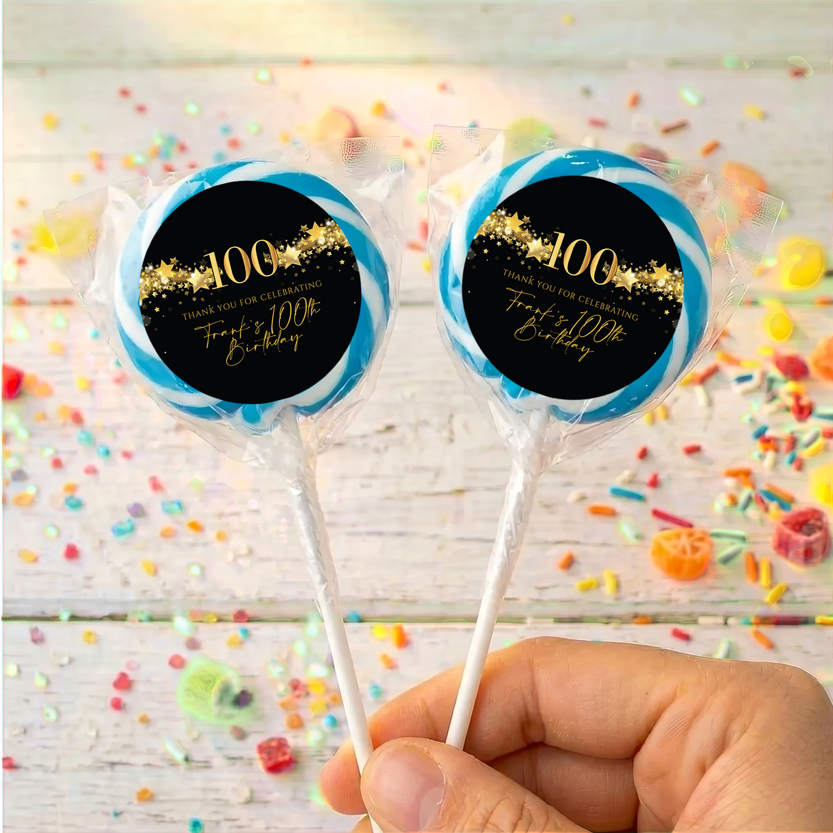 Two 100th birthday lollipops with black gold star labels and blue rim, held up