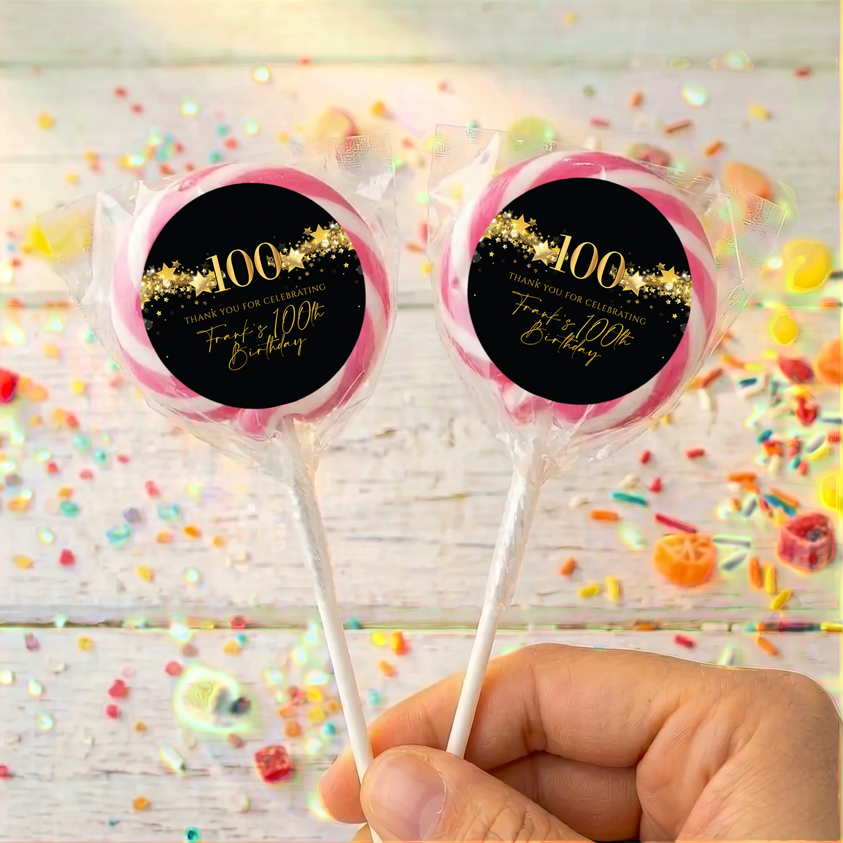 Two 100th birthday lollipops with black round gold star labels, pink rim, held