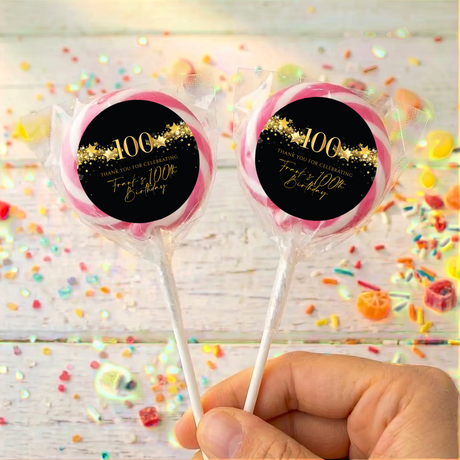 Two 100th birthday lollipops with black round gold star labels, pink rim, held