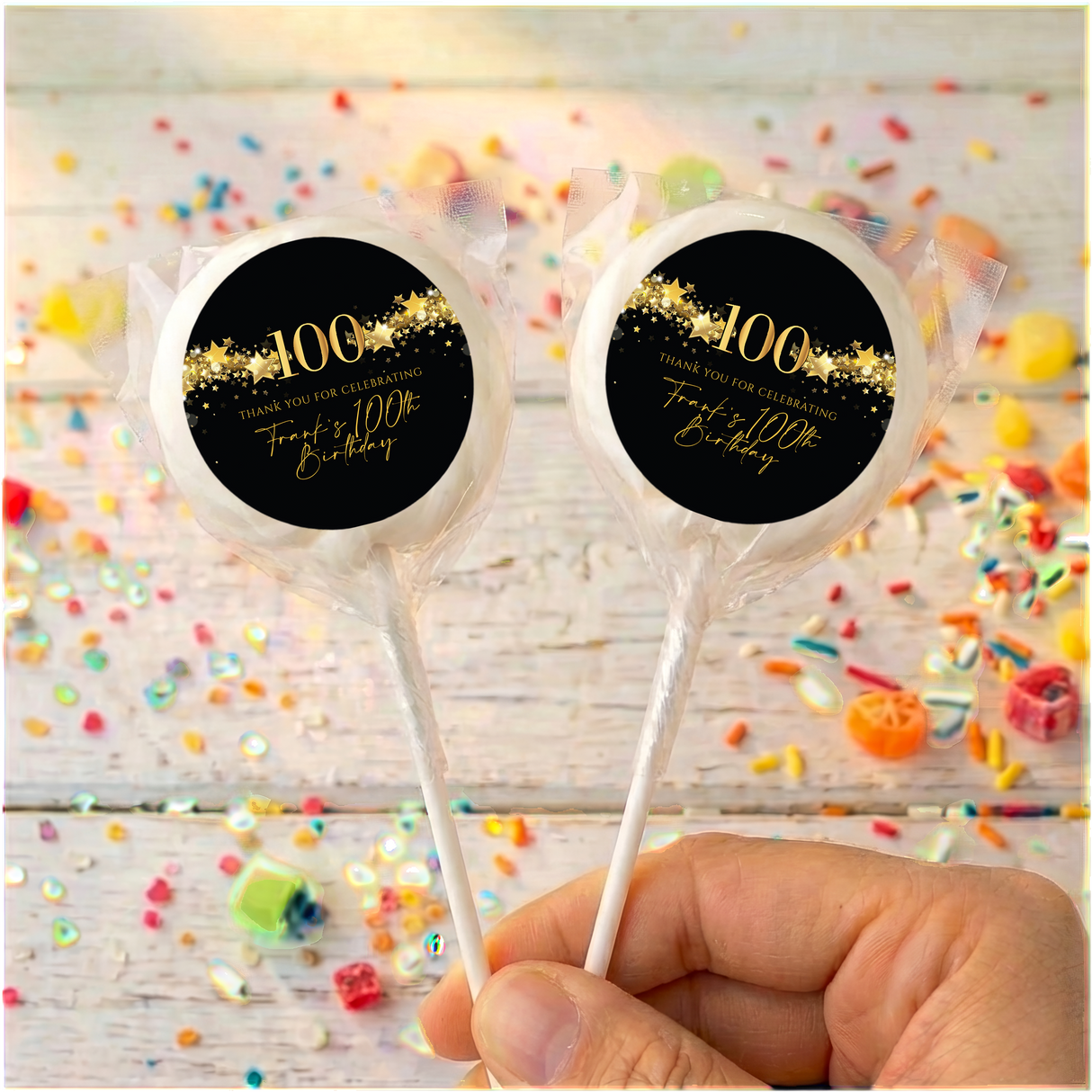 Two 100th birthday lollipops with black gold star labels in clear wrappers, held up