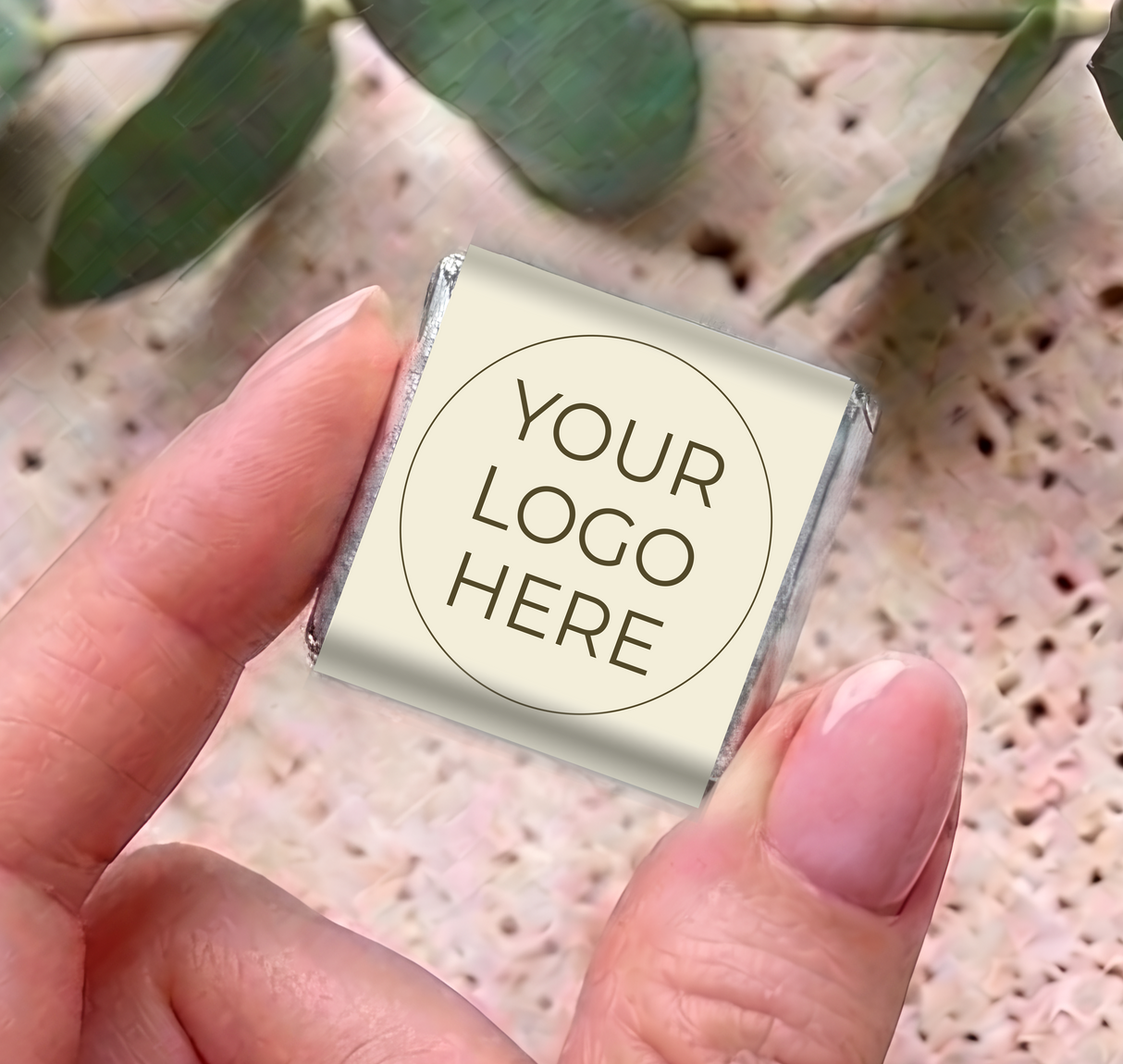 Mini personalized chocolates with “YOUR LOGO HERE” on cream wrapper and silver foil ends, held