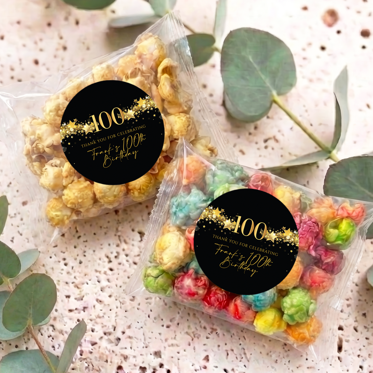 Two personalised 100th birthday popcorn bags with clear wrappers and black-gold round stickers.